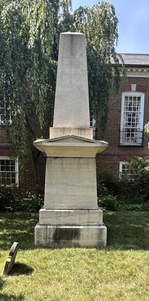 Tall white obelisk monument for Charles Bennett standing on a stepped pedestal in the Christ Church churchyard, with the brick church building and trees visible behind it.