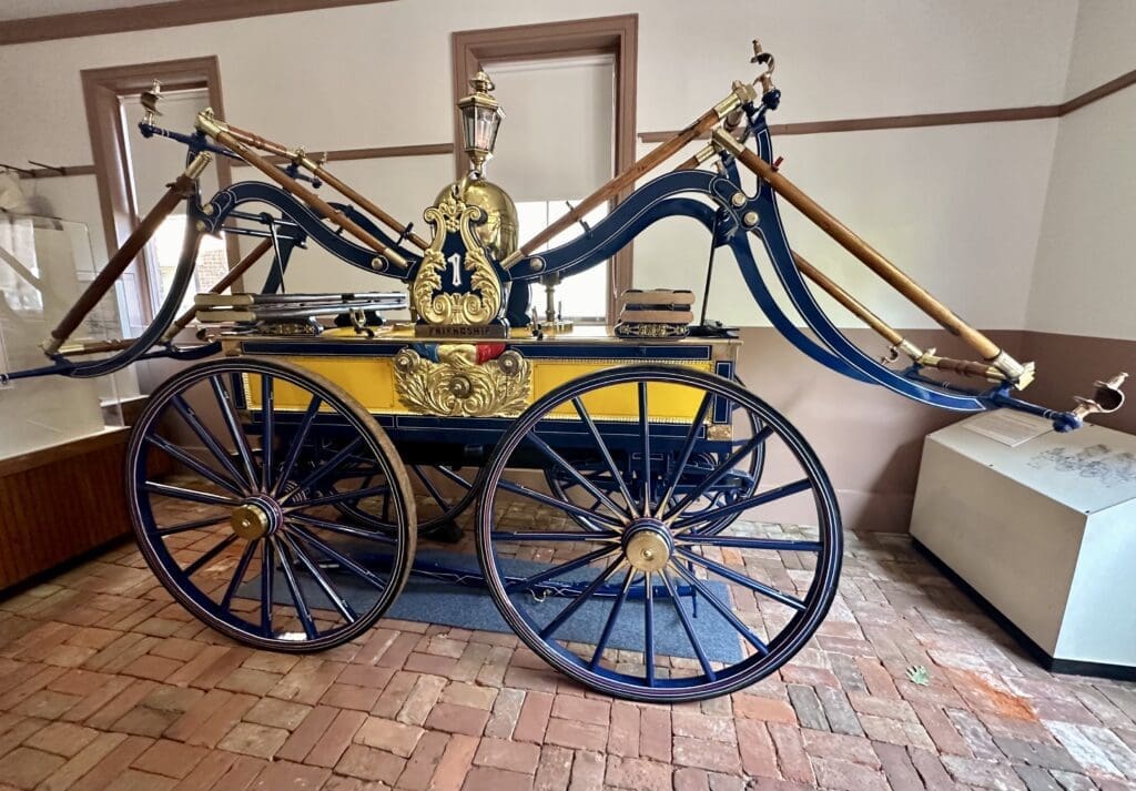 Restored 1851 suction engine with large wooden wheels and brass fittings, displayed inside the Friendship Firehouse Museum in Alexandria; involved in the 1852 fatal accident of Charles Glasscock.