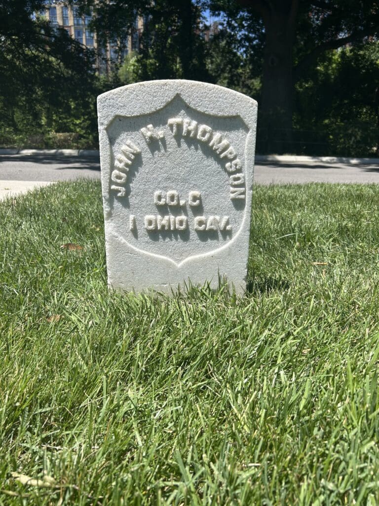White, shield-shaped cemetery headstone standing in a grassy lawn with trees and a road in the background