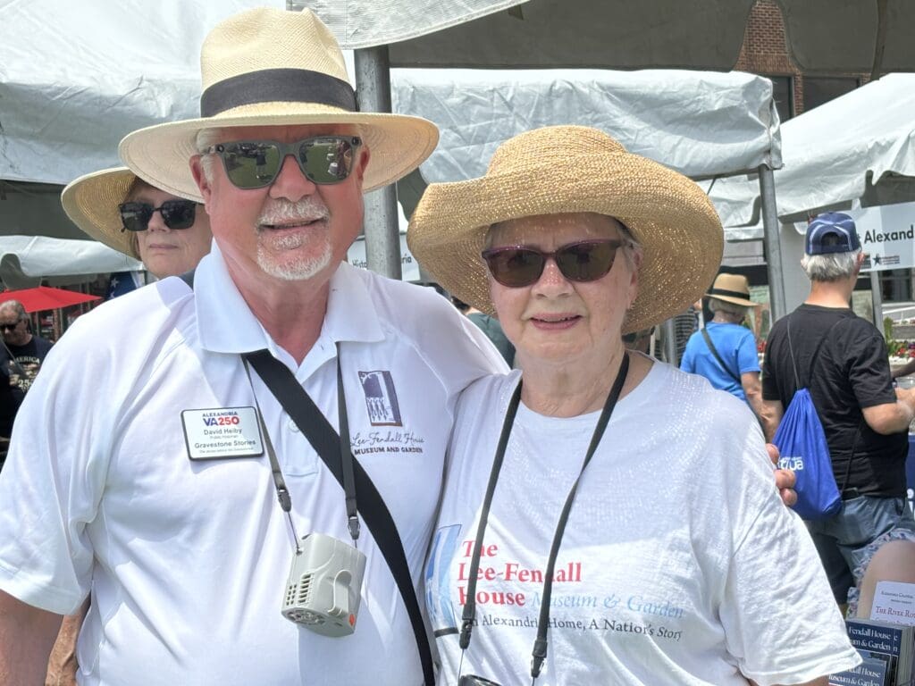 Public historian David Heiby and his wife, Christine, smiling together beneath a tent at the Lee-Fendall House Museum booth during Sails on the Potomac on the Alexandria waterfront. Both wear straw sun hats, sunglasses, and white Lee-Fendall House Museum and Garden shirts; Heiby wears a VA250 name badge identifying him as a public historian with Gravestone Stories.