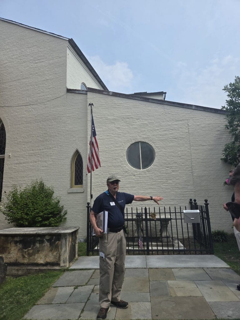 David Heiby gestures beside the fenced Tomb of the Unknown Soldier, with an American flag and the Meeting House behind him.
