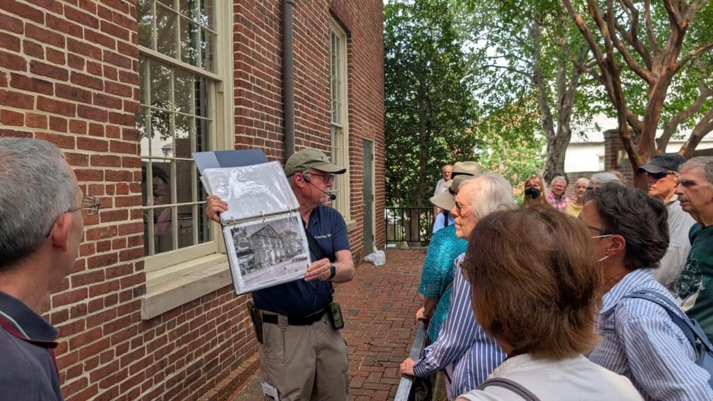 David Heiby holds open a binder of historic photographs while speaking to tour participants beside the brick Meeting House.