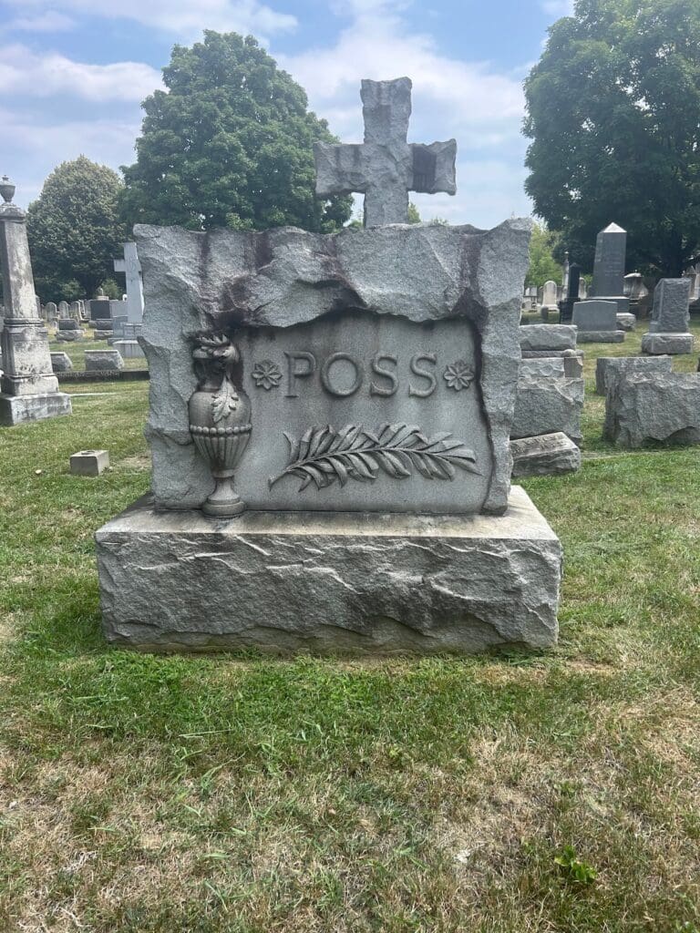 Gravestone with a cross on top, carved with the name P.O.S.S. and a decorative vase and laurel motif in a cemetery setting