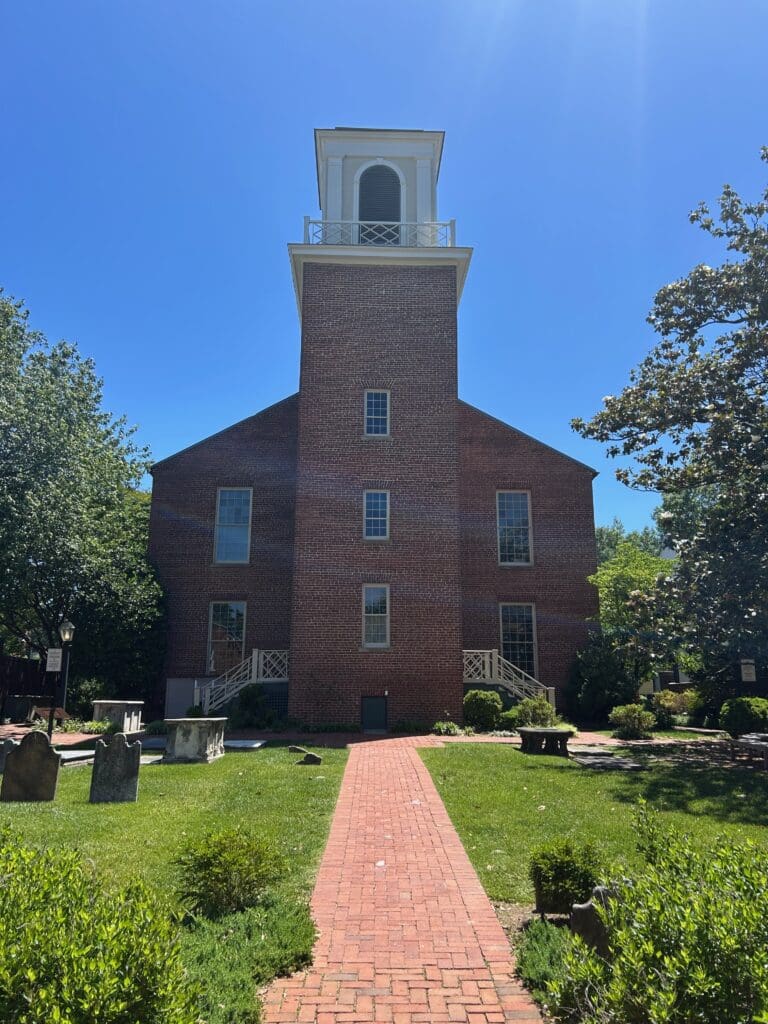 Brick bell tower and south wall of the Old Presbyterian Meeting House in Alexandria, Virginia, seen across the burial ground, with 18th-century headstones and a raised table stone in the grass and a brick walkway leading to the tower door.