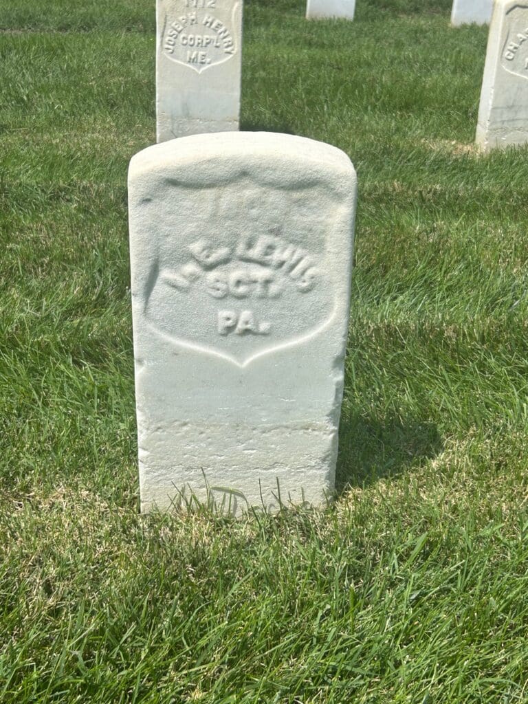 Weathered white marble Civil War headstone of Sergeant Isaac E. Lewis of Pennsylvania, Grave 1600, at Alexandria National Cemetery, with other military headstones visible behind it.