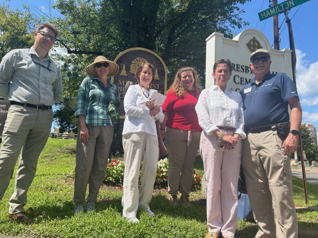 Six people stand smiling on a grassy rise inside the Wilkes Street Cemetery Complex on a sunny day, flanked by two cemetery entrance signs — the wooden Methodist Protestant Cemetery marker and the white Presbyterian Cemetery sign — with a green Hamilton street sign and headstones visible behind them.