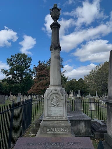 Obelisk gravestone of Francis Lee Smith in the Presbyterian Cemetery, Alexandria, Virginia, topped with a draped urn symbolizing mourning and eternal life, with the cemetery's wrought iron fence visible behind it.