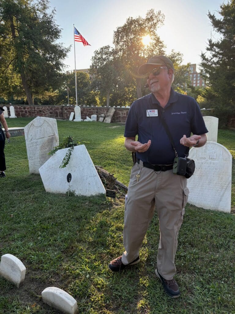 Public historian David Heiby, in a Gravestone Stories polo and headset, speaks with open hands beside the marked grave of Revolutionary patriot Abraham Faw in Trinity Methodist Church Cemetery, Alexandria, with an American flag and rows of headstones behind him in late-afternoon sun.