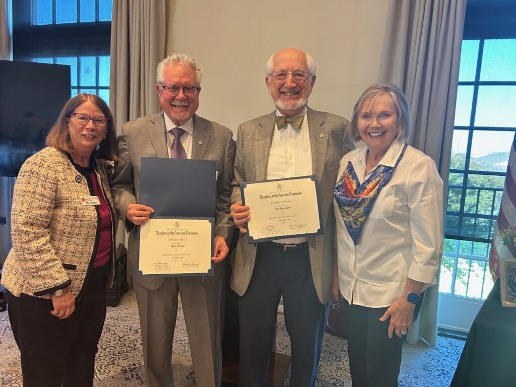 Four people smiling in a sunlit room with tall windows at the Belle Haven Country Club. From left: Meg Kane, Regent of the Kate Waller Barrett Chapter, NSDAR; David Heiby and Don Dahmann, each holding a Daughters of the American Revolution Certificate of Award for Excellence in Community Service; and chapter member Sandy Welch.